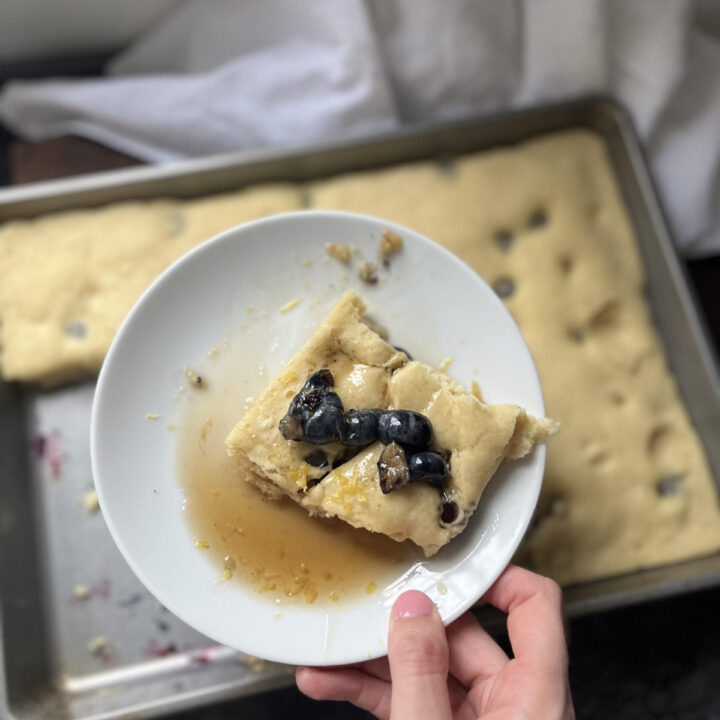 Hand holding plate with blueberry lemon pancake with syrup on top and more pancakes in background