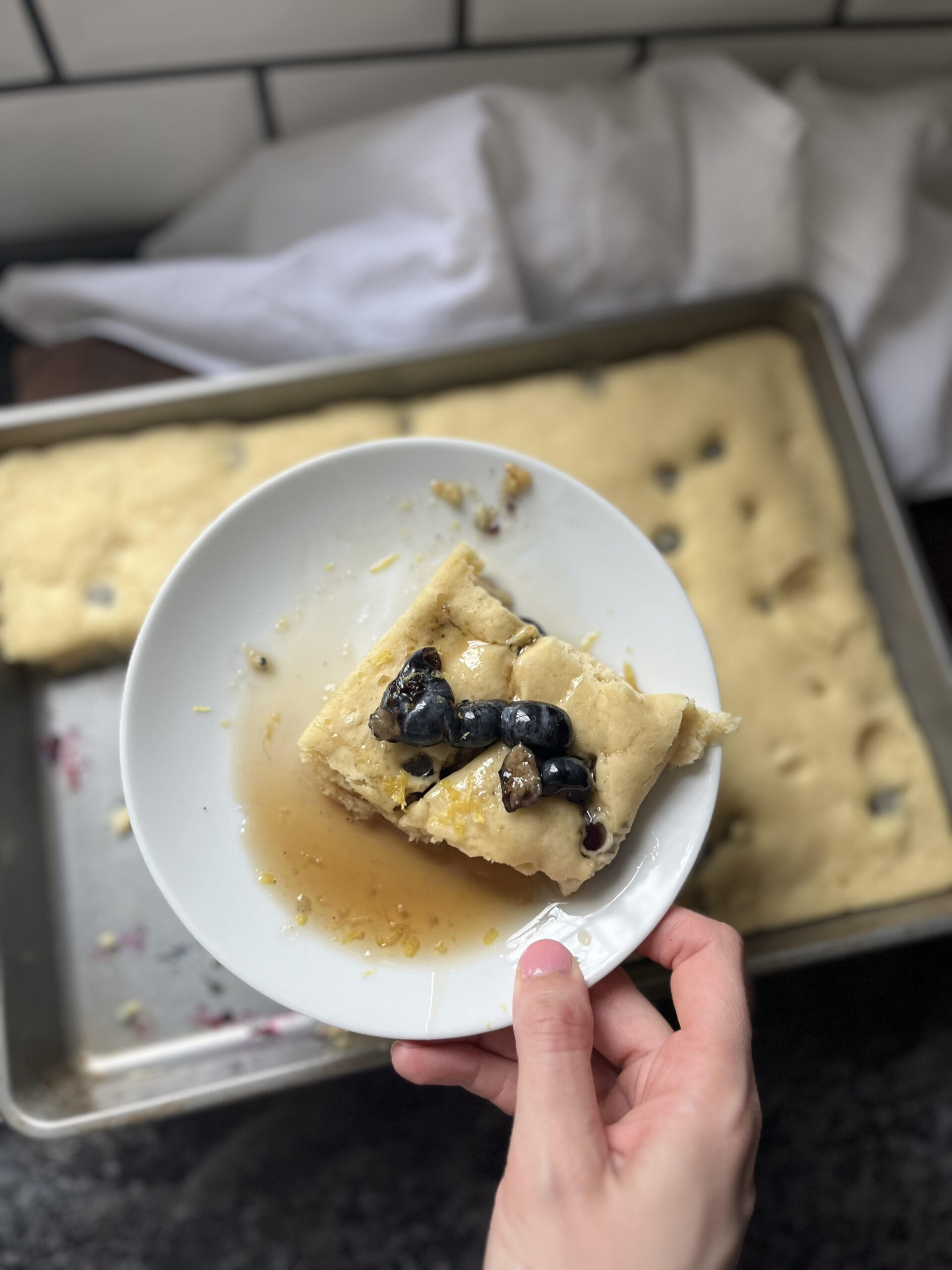 Hand holding plate with blueberry lemon pancake with syrup on top and more pancakes in background