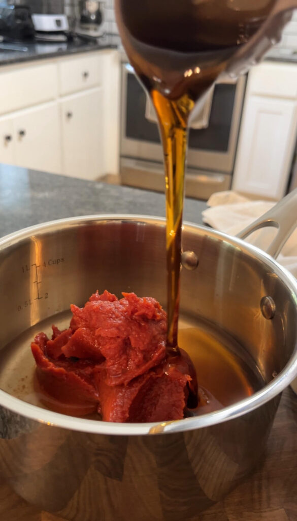 Measuring cup drizzling raw blue agave into homemade ketchup in a stainless steel pot